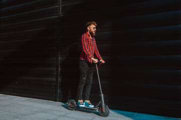 Fototapeta na wymiar Full lengh of handsome young bearded caucasian businessman rides a scooter in front of a black wall