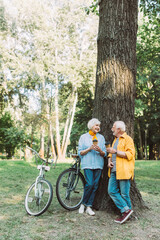 Elderly couple smiling at each other while holding paper cups near tree and bikes in park
