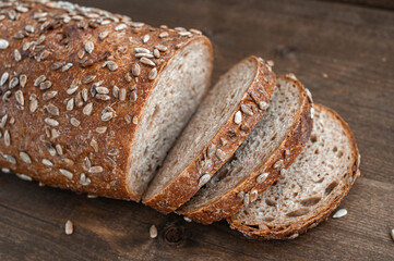 A loaf of fresh wholegrain bread cut into pieces on a brown wooden background.