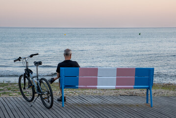 Older person with bicycle on a bench painted in the representative colors of transsexuality.