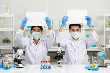 Two Male Scientists wear Face Mask in Lab while holding blank paper. Copy Space. SARS-CoV-2 , Covid-19 THEME.