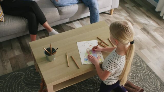 Small girl stays home, busy with drawing beautiful picture of happy family, lacking communication with parents that are sitting on sofa, sucked by Internet, Top view, Slow motion.