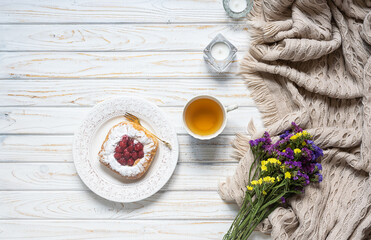 Romantic composition with warm blanket, cup of herbal tea, candles and tasty homemade cake on white wooden background. Hygge home interior. Lagom concept.