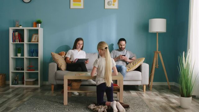 Young parents, mom and dad, are sitting on sofa in living room, spending time online, using gadgets, ignoring small daughter that is drawing in front of them, concept of generation gap, Slow motion.