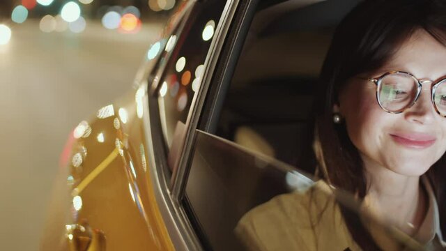 View from outside of the car of beautiful brunette woman looking at evening city through the window and smiling while riding in the backseat