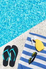 Flip flops, sunscreen spray and sunglasses on a blue and white striped towel at the edge of a pool....
