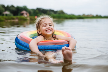 Fototapeta premium Happy child swims in the river on a colored inflatable circle. Cute little girl gives birth while swimming in the lake. Summer vacation.
