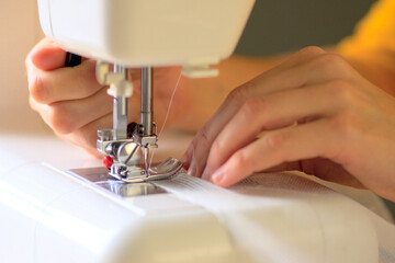 The process of sewing curtains from a mesh of white fabric. The rolled edge of the fabric is placed under the presser foot of the sewing machine. Close up view.