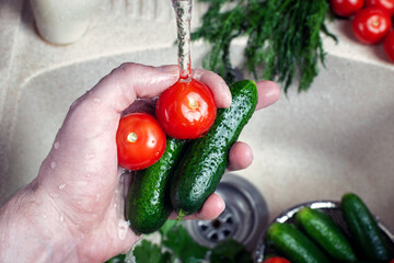 close-up of a man's hand holding several fresh cucumbers and tomatoes under the tap water in the sink, health and hygiene of washing vegetables