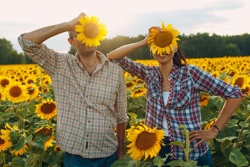 Man and woman farmers at sunflower field holding flower on head.