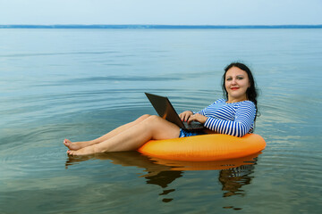 Woman on vacation works remotely with a laptop in a swimming circle in the morning water.