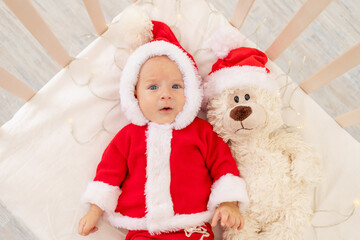 Christmas photo of a baby in a Santa costume lying in a crib at home with a toy in a Santa Claus hat, top view, happy new year