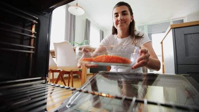 culinary, food and people concept - woman cooking salmon fish in baking dish in oven at home kitchen