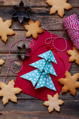 Handmade New Year origami postcard with paper christmas fir tree. Holiday idea for family time and gift for beloved. Gingerbread cookies and molds, wooden background. Flat lay top view