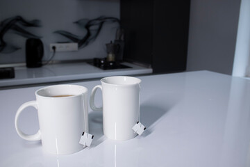 two white cups with tea and sticking out tea bags on a white table in a gray kitchen. High quality photo
