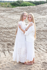 Fototapeta na wymiar young pretty twins with long blond hair hold hands at sand quarry in elegant white dress, skirt, jacket. stylish fashion photoshoot, summer photosession. identical sisters spend time together outdoors