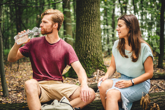 Caucasian couple stopped to rest while walking in the forest, young people sit on a fallen tree trunk drinking water from a bottle. Active rest, hike in a wooded area. Thirst and water balance