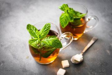 Pfefferminztee in zwei Gläsern, Löffel und Würfelzucker auf einem grauen Hintergrund. Nahaufnahme, frische Minze, Teepause, rustikaler Stil.