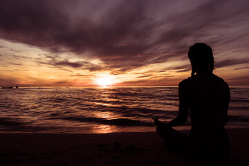 Silhouette of young woman wearing exercise clothes practicing yoga on the beach at sunset or sunrise. women do yoga. women exercising yoga in the beach.Young girl meditating in lotus posture.