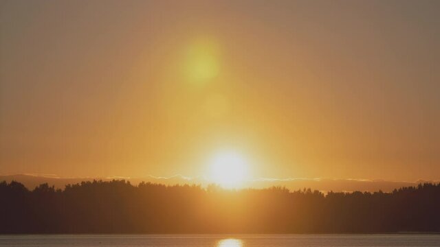 Time-lapse sunrise over beautiful lake. Water surface in fast motion. Warm sky with rolling clouds. Trees silhouette woodland on horizon. Early morning shot in timelapse. Bright sun rises over skyline