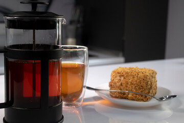 cup of tea in a double-botoom glass, french-press teapot and piece of cake on white table in gray kitchen. High quality photo