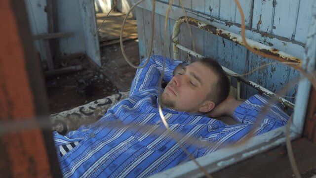A young man in pajamas is lying on an old bed in a dirty, dilapidated house