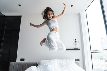 Image of young excited woman making fun and jumping on bed