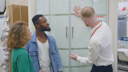 Portrait of happy multiethnic couple standing with assistant near new shower cabin in modern shop