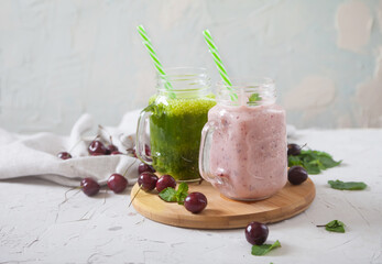 Green and sweet cherry smoothies on a wooden stand, scattered cherries and a linen napkin on a concrete background.