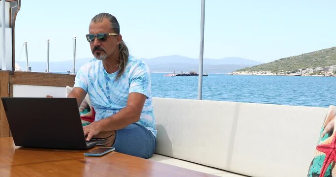 Portrait of a middle aged freelancer man is sitting on the deck of a yacht with a laptop computer at summer vacation