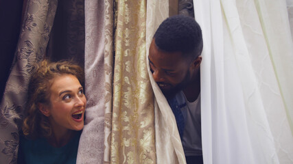 Happy diverse couple shopping in textile store having fun and hiding in curtains