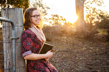 Happy young writer holding pen and notebook leaning on fence post in paddock