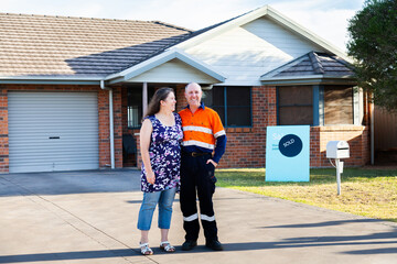 Happy middle aged couple standing infront of house with sold real estate sign