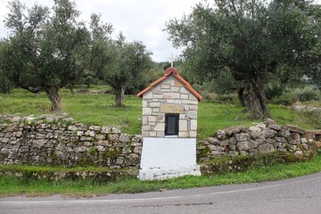 Obraz premium A small country iconostasis, church-like small-scale replica for candle-lighting with olive grove in the background in Messinia region, southwestern Greece.