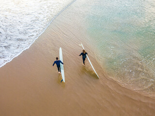 Looking down on two surfers walking on a beach between waves