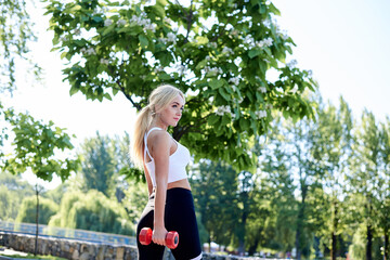 Young blond woman, wearing white top and black leggings,,doing power morning exercises with red fitness weights in city park in summer.Training process outdoors. Active healthy lifestyle concept