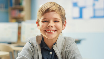 Portrait of a Cute Little Boy Sitting at his School Desk, Laughs Happily. Smart Little Boy with Charming Smile Sitting in the Classroom. - obrazy, fototapety, plakaty