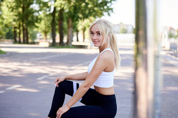 Young fit blond woman, wearing white top and black leggings, squatting down, posing by city lake in park in summer. Sportswoman, resting relaxing after fitness exercise training. Healthy lifestyle.