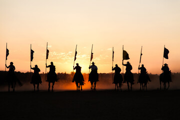 Line of silhouetted horses and riders with flag poles charging at sunset