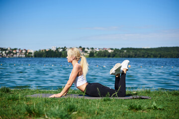 Young blond woman, wearing black leggings and white top, doing stretching exercises for back on grey yoga mat by lake in summer morning. Female sport training outside on fresh air. Healthy lifestyle.