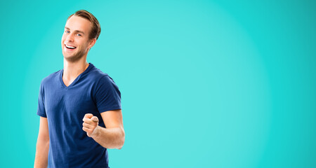 Happy smiling handsome man in clothing, showing copy space, visual imaginary or something, or pressing virtual button, aqua marine blue color background. Pointing at You - advertising concept picture.