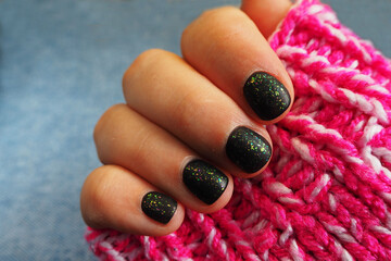 a woman's hand with nails is covered with black gel Polish with green sequins and a matte effect on the background of a pink knitted sweater . warmth and comfort