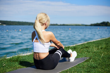 Young blond woman, wearing black leggings and white top, stretching back and legs on grey yoga mat by city lake in summer morning. Female sport training outside on fresh air. Healthy lifestyle.