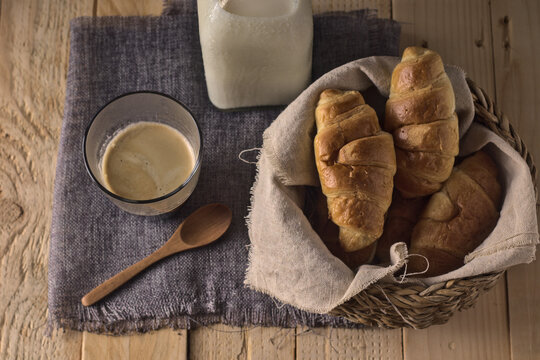 croissant reci&eacute;n horneados sobre un cesto de mimbre en una mesa con caf&eacute; y leche, listo para tomar en desayuno o merienda