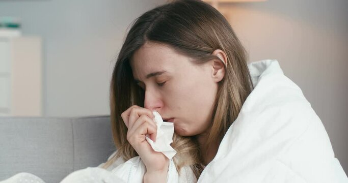 Close up of sick young bad-looking Caucasian woman in blanket sitting at home on couch, sneezing, caughing and blowing nose in napkin. Coronavirus concept. Female with bad sympthoms of flu.