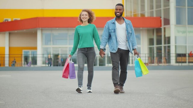 Young diverse couple holding hands when walking outdoors with paper bags after shopping