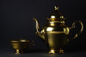 Gold tea set on a black background. The teapot and teacup are next to each other.