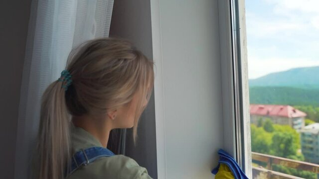 The blond girl in the yellow gloves is washing a dirty window with a blue cloth