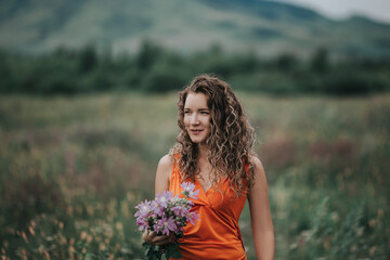 A girl in an orange dress with a bouquet of flowers walking along the field against the background of grass and mountains