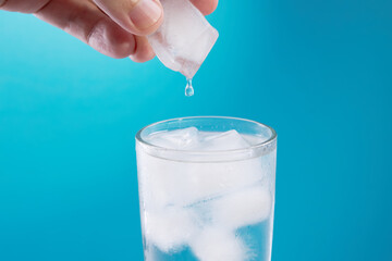 person putting ice to water glass close up view over blue background. copy space. refreshment beverage. healthy lifestyle concept